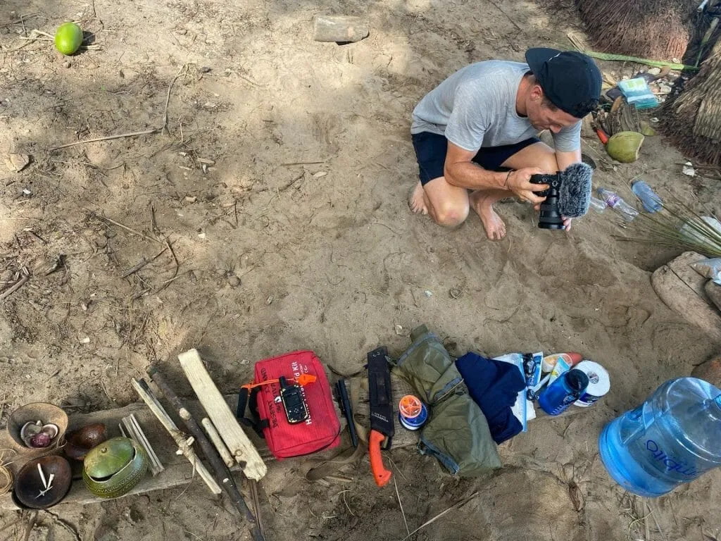 Kara and Nate behind the scenes: Man filming survival gear on a sandy island.