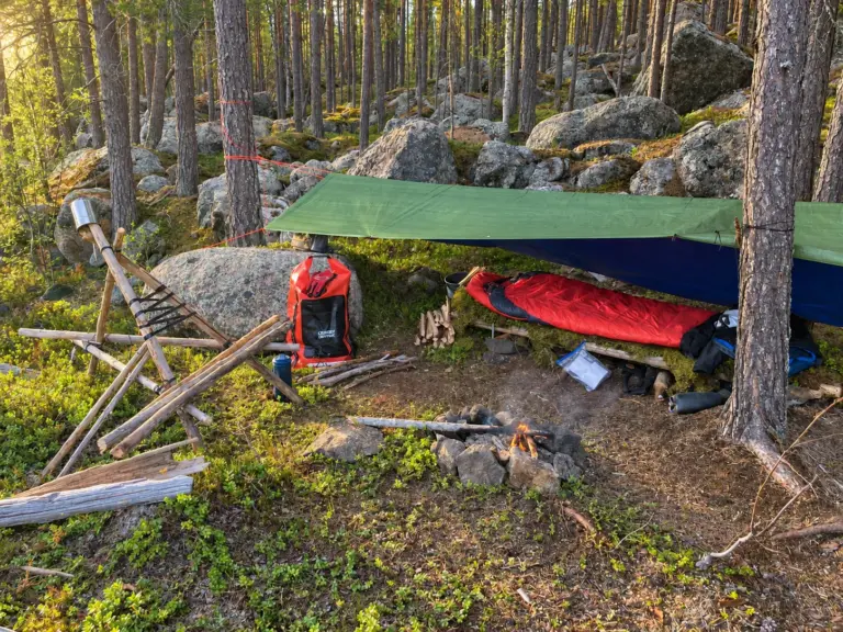 Solo wilderness camp with a green tarp, red sleeping bag and dry bag among pine trees and boulders