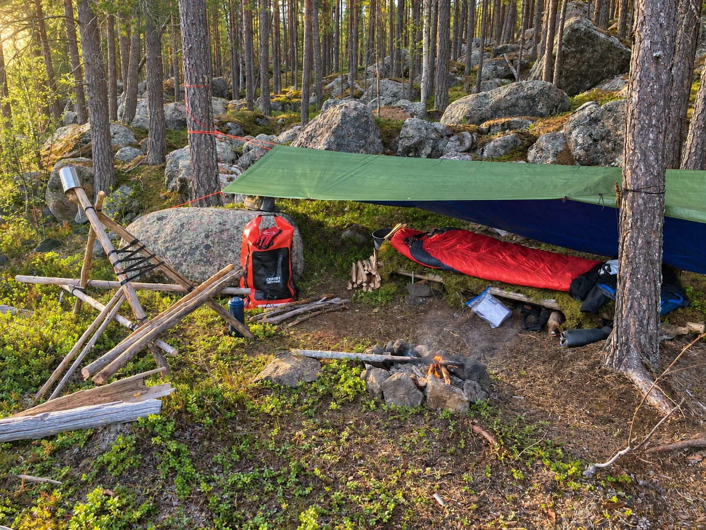 Solo wilderness camp with a green tarp, red sleeping bag and dry bag among pine trees and boulders