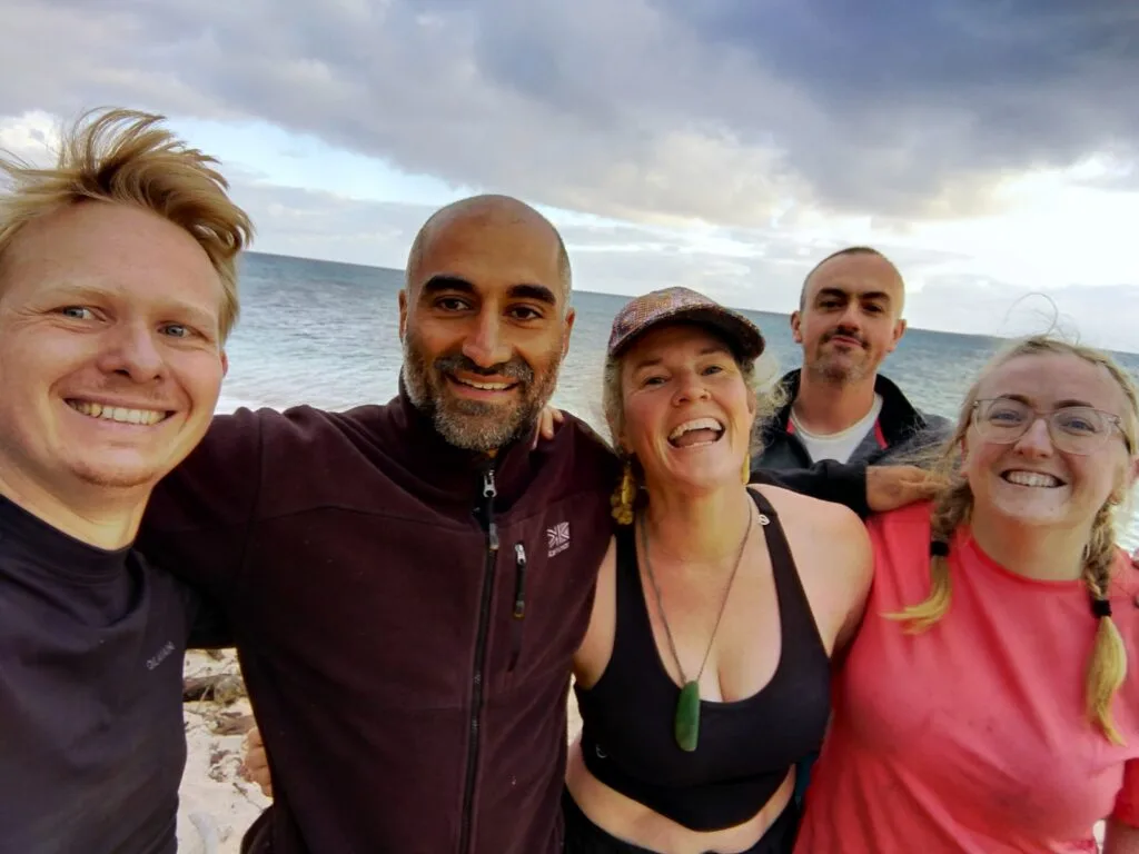 Five smiling adults stand close together on a beach with the ocean and cloudy sky in the background, appearing happy and.