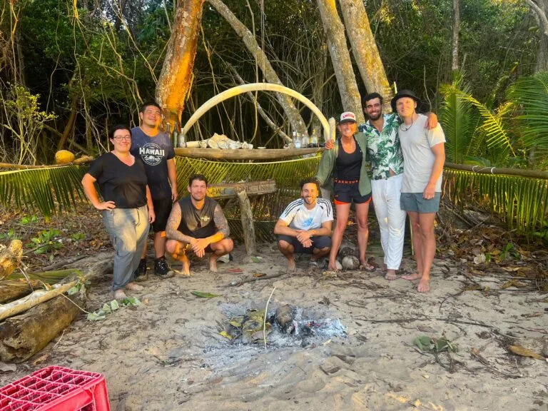 Seven people stand and kneel on a sandy beach in front of a campfire, surrounded by trees and greenery. A makeshift table.