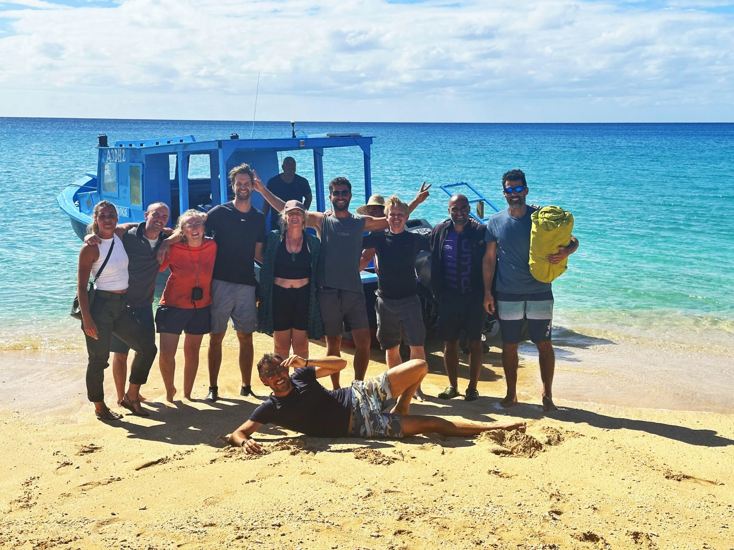 A group of eleven people smile and pose together on a sandy beach with clear blue water and a blue boat in the background.