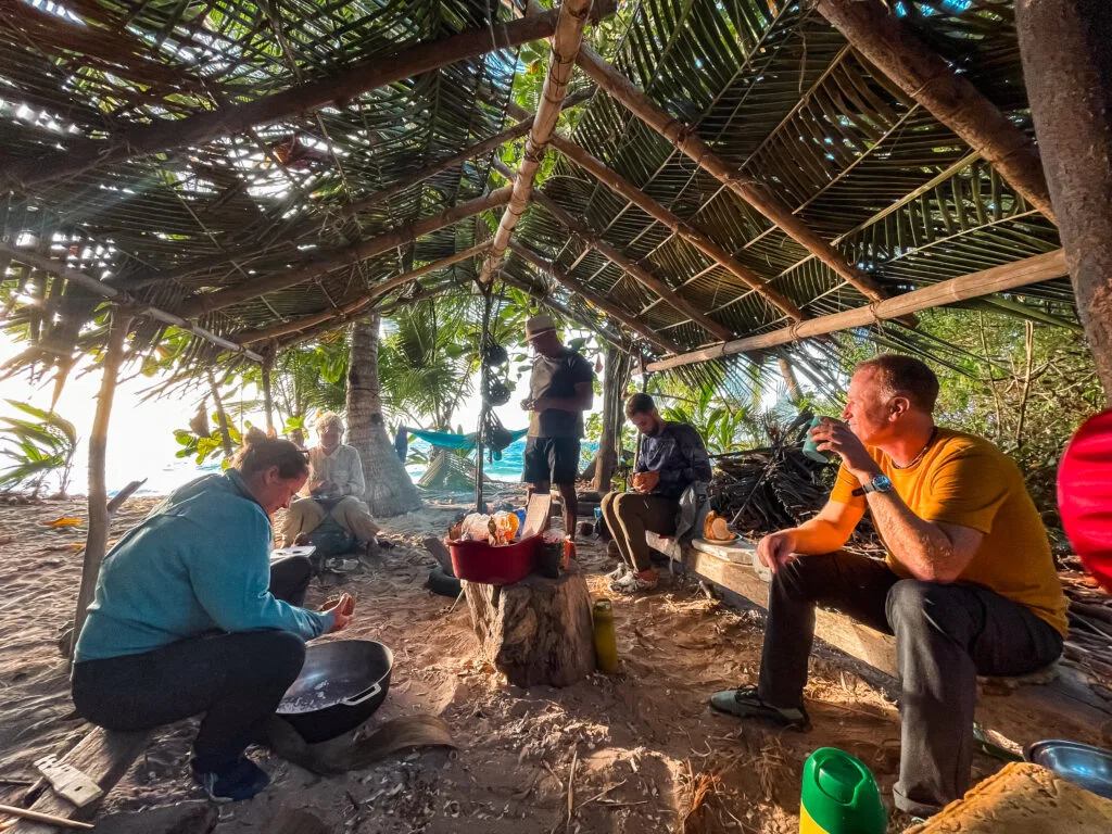 A group of people are enjoying a meal under a palm frond-thatched shelter on a beach.  Several individuals, likely researchers or travelers, are preparing and eating a meal around a small fire.  The scene suggests a relaxed, informal gathering, perhaps a break from fieldwork or exploration.  The natural setting and the simple shelter highlight a connection with nature.
