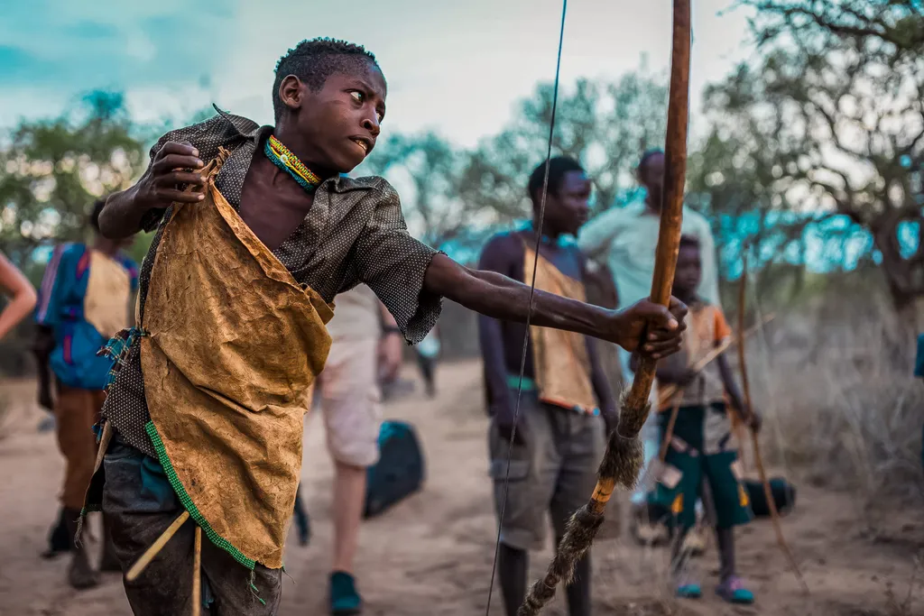 A boy in traditional clothing aims a bow and arrow outdoors during the Hadza Tribal Bushcraft Experience & Luxury Safari, surrounded by children and adults in a scenic rural setting.