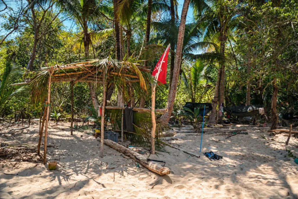 Simple hand-built palm-leaf shelter at a beach camp, with hammocks strung in the trees behind