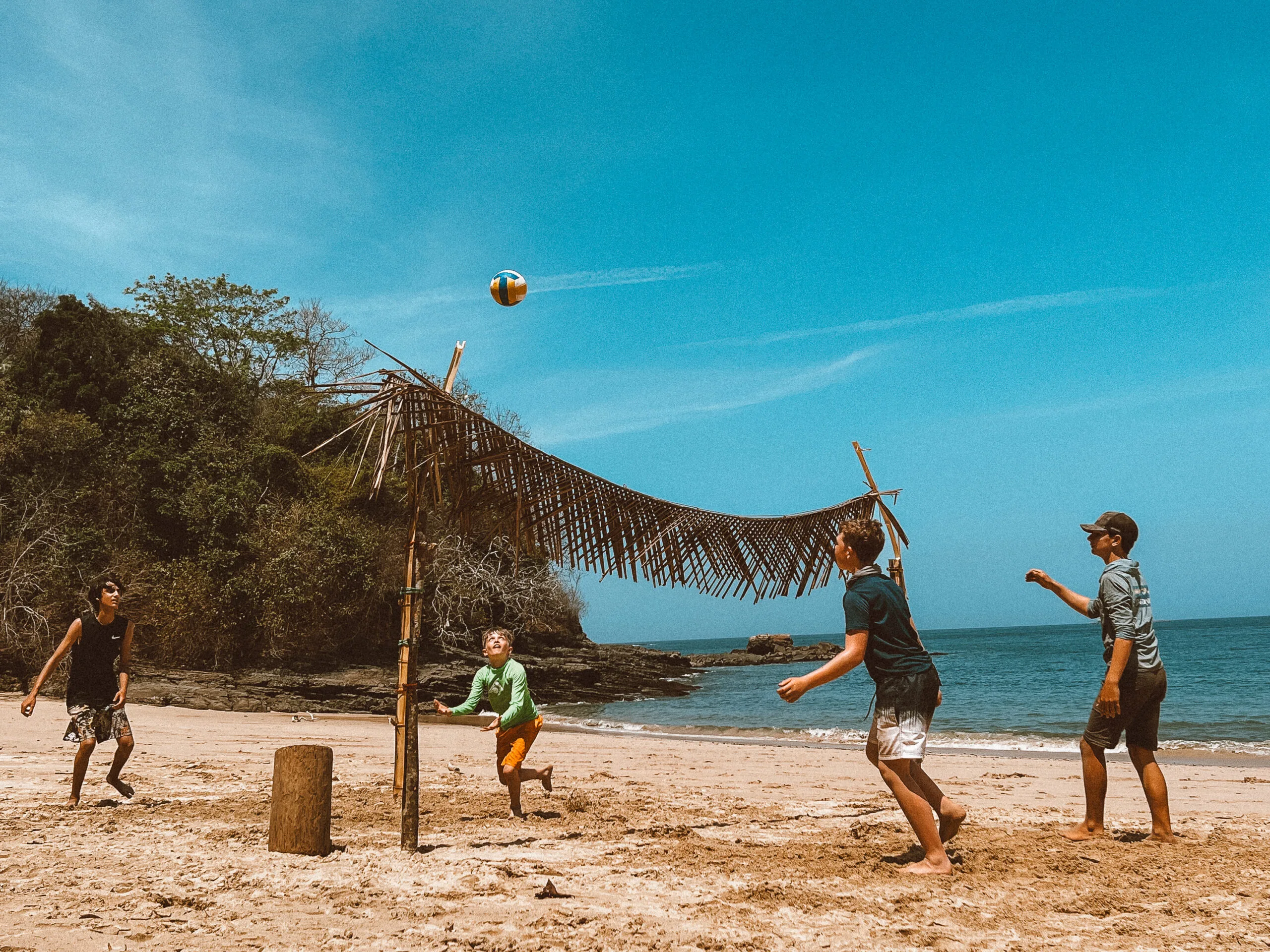 Kids playing beach volleyball on a tropical desert island with a makeshift palm leaf shade and ocean in the background.