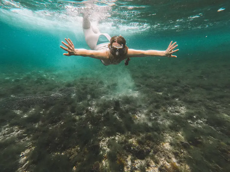 A person wearing goggles and a snorkel swims underwater above a seabed covered in algae and rocks, arms extended forward.
