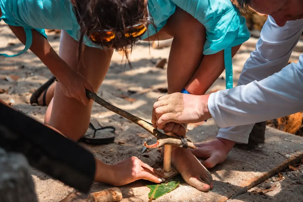 Two participants working a bow drill together to light a friction fire on the beach