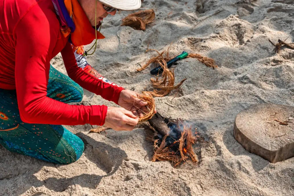 Participant nursing a coconut-husk tinder bundle into flame on the sand