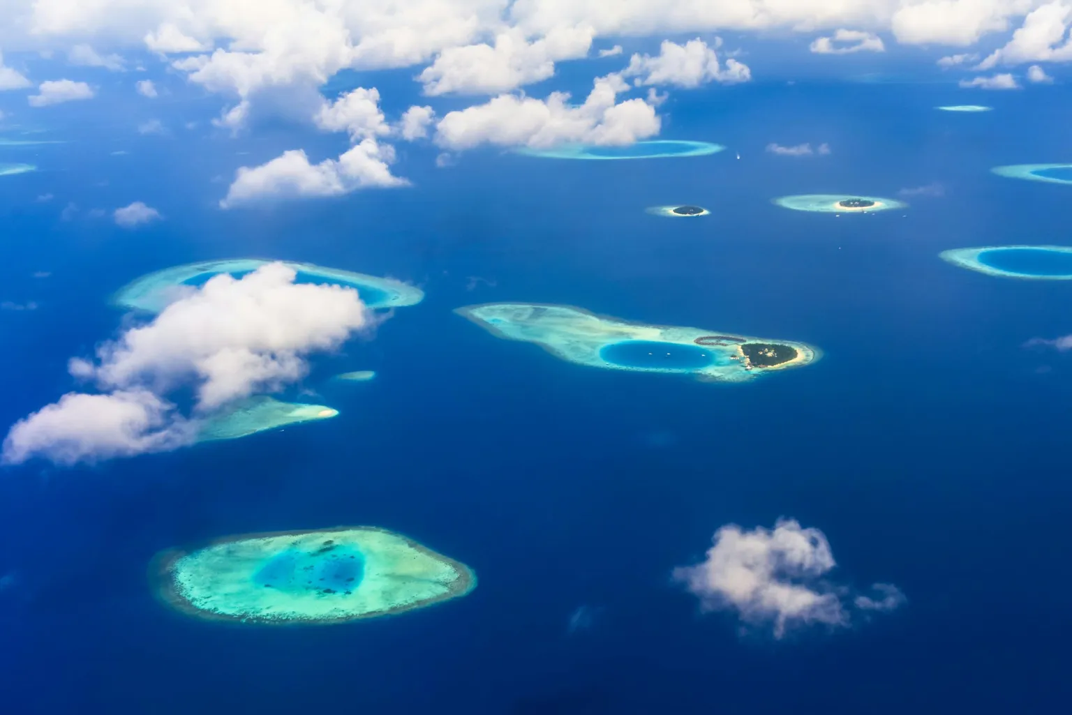 Aerial view of turquoise atolls surrounded by deep blue ocean and scattered clouds, capturing the essence of an Island Survival Adventure in the Maldives.