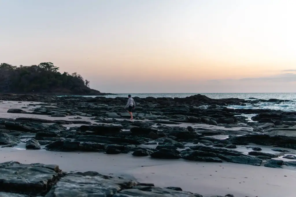Secluded desert island beach at sunset with rocky shoreline and a person exploring the coast, emphasizing survival and adventure in remote island environments.