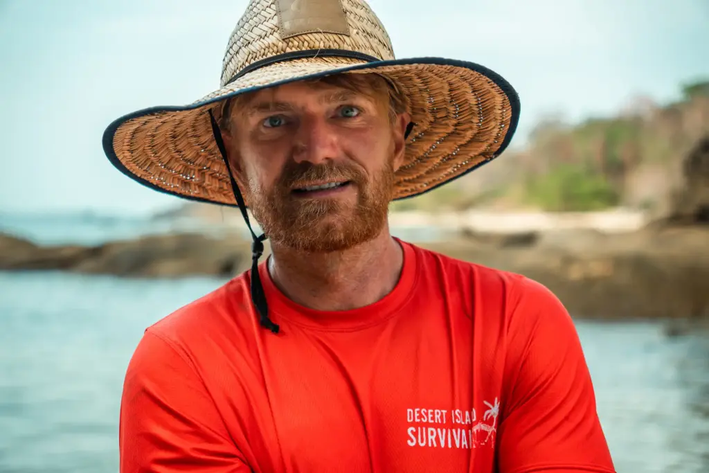 Man wearing a wide-brimmed straw hat on a deserted island beach for survival tips.