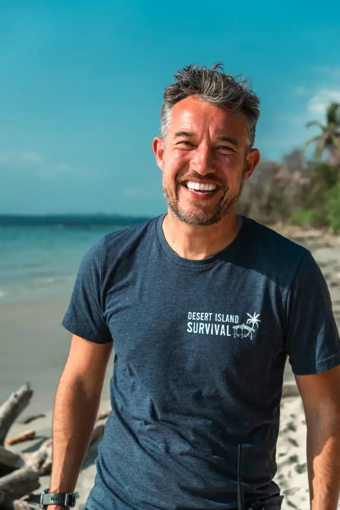Survival expert on tropical beach with "Desert Island Survival" shirt, coastal scenery, sunny weather.