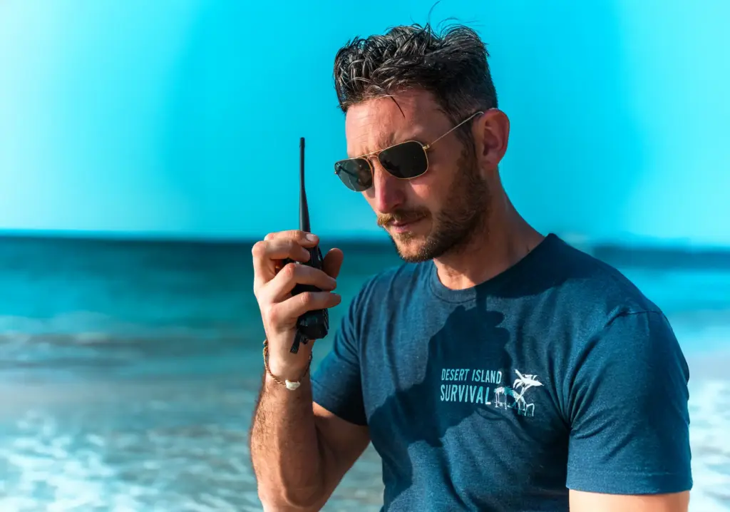 A man wearing sunglasses, holding a walkie-talkie, at a beach, promoting desert island survival skills.