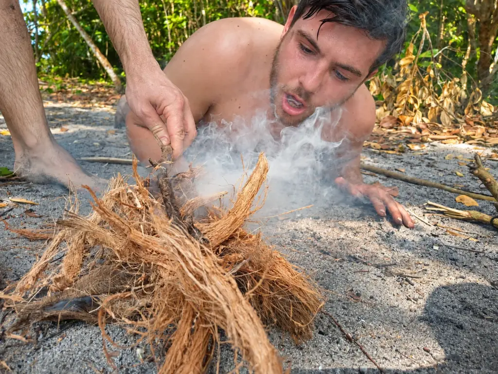 Participant blowing a smoking coconut-husk tinder bundle into flame during friction fire practice