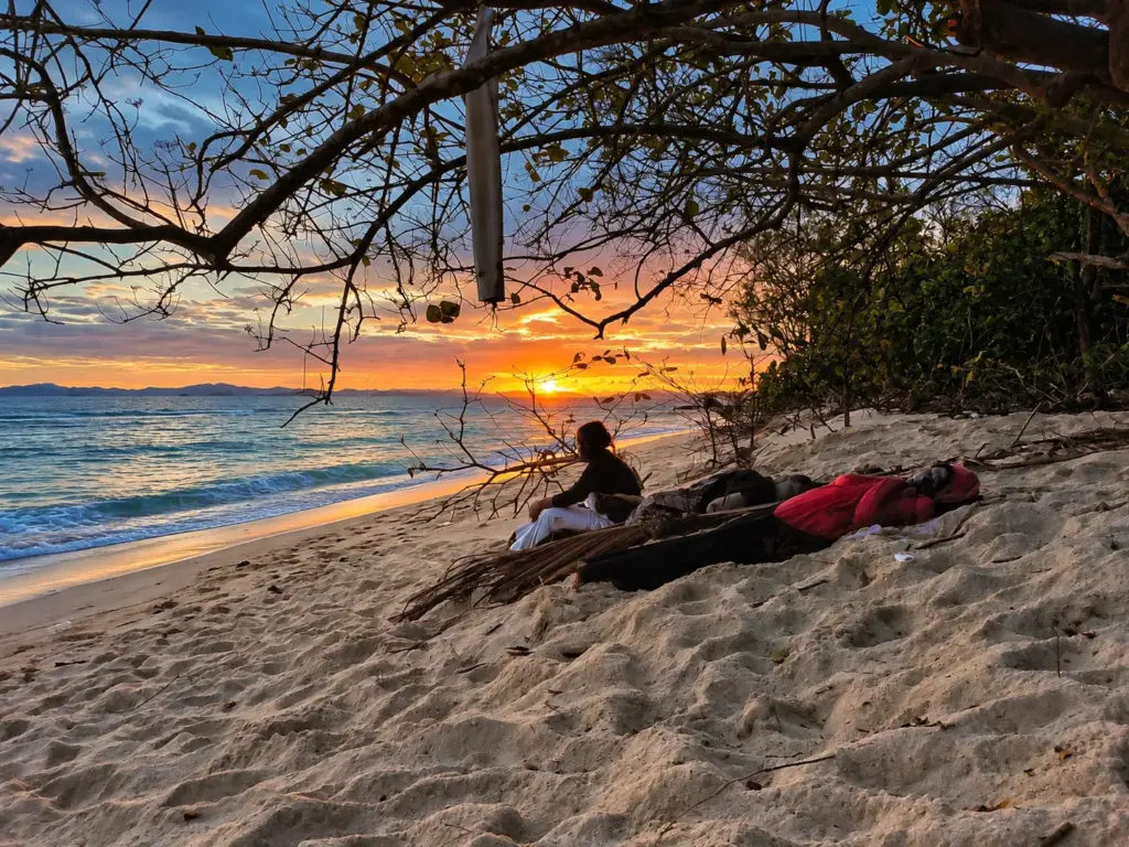 Person resting on a quiet island beach at sunrise beneath overhanging branches