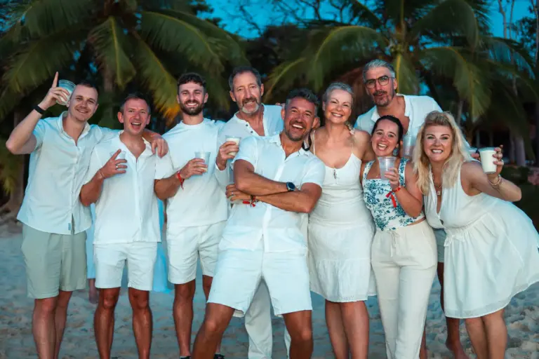 Smiling group of friends enjoying a tropical beach party on a desert island with palm trees at sunset, celebrating.