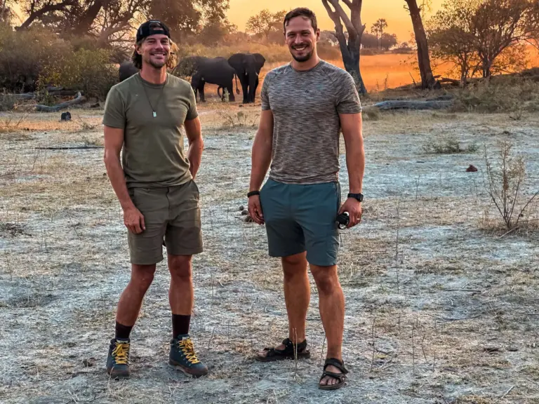 Men standing on dry savannah with elephants in the background during sunset, illustrating adventure and wildlife experience.
