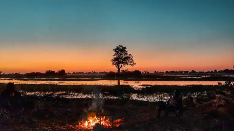 Serene sunset over a desert island with a lone tree, reflecting in calm waters, and two people sitting by a campfire.