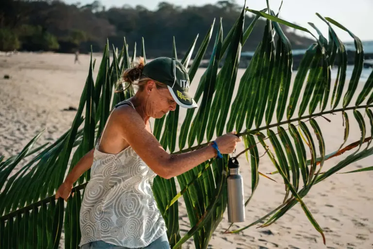 Lush green palm leaves on a sandy beach with a woman in casual clothing and a cap, holding a water bottle and exploring.