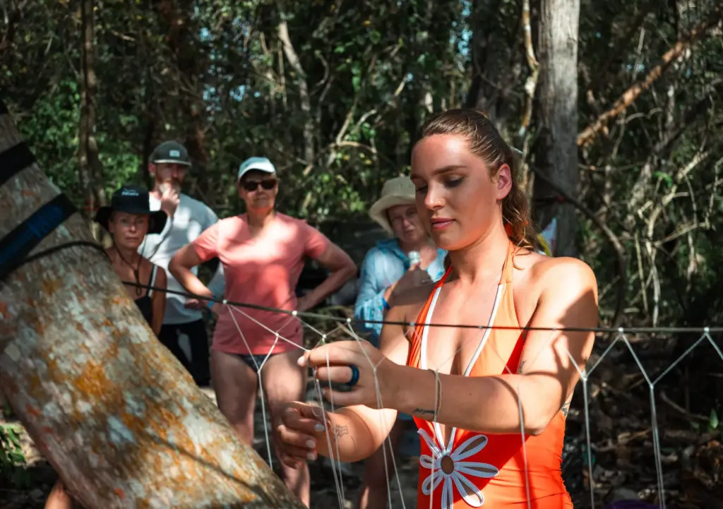Woman demonstrating bushcraft skills, tying knots with a group watching in a forest setting.