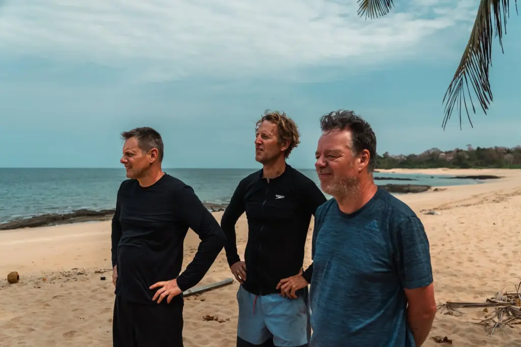 Men standing on tropical desert island beach, contemplating survival strategies, with ocean background and tropical palm leaves, emphasizing desert island survival skills and adventure in a remote location.
