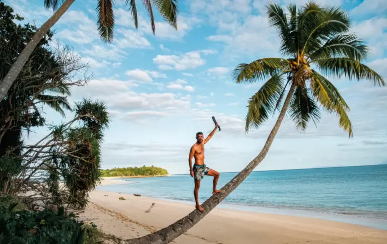 A man climbing a leaning palm tree on a deserted tropical beach, with clear blue sky and calm ocean, showcasing survival.