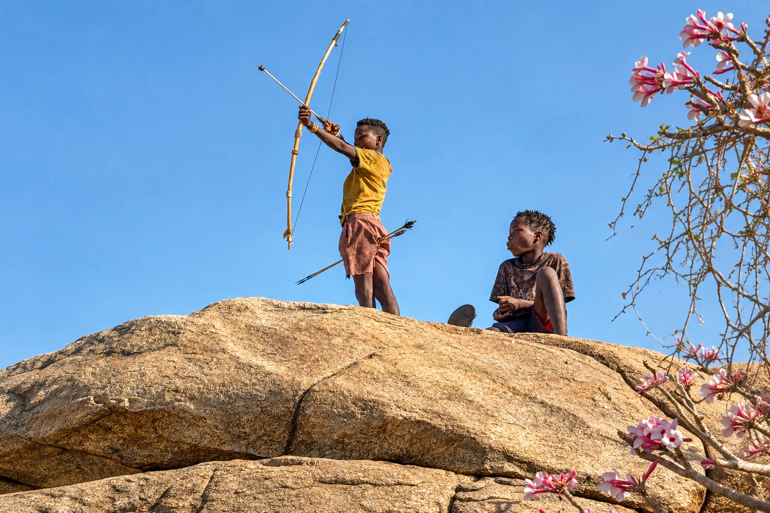 Young Maasai boy aiming a bow and arrow with another boy watching on a rocky outcrop in Tanzania.