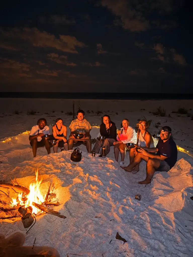 The group sits in a circle around the beach campfire after dark