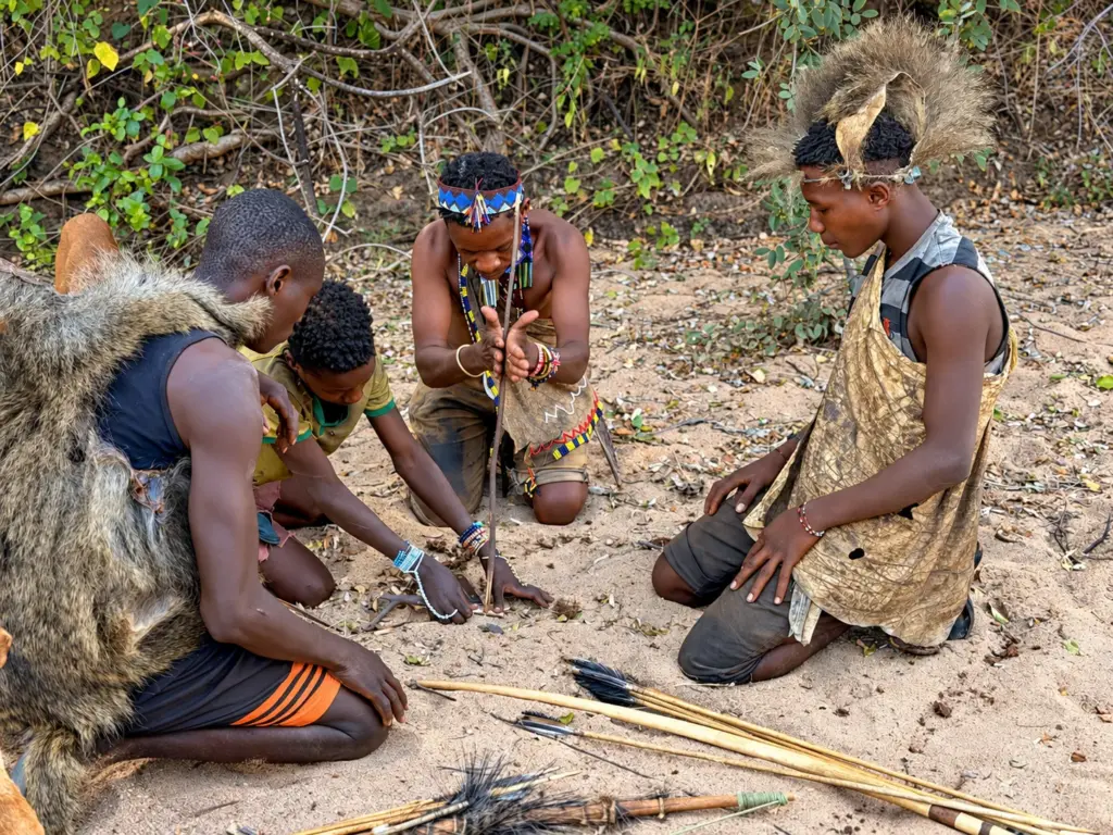 Hadza men kneel on the sand making fire with a hand drill, their bows and arrows laid beside them