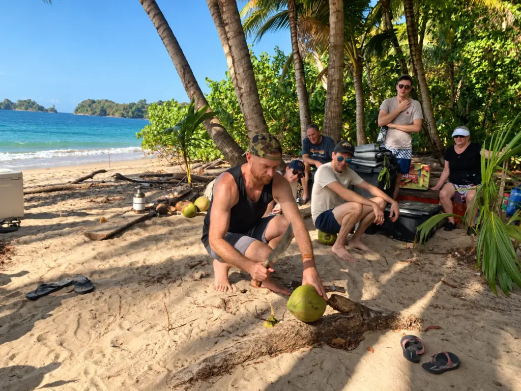 Participants rest in the shade of their beach camp while one opens a coconut