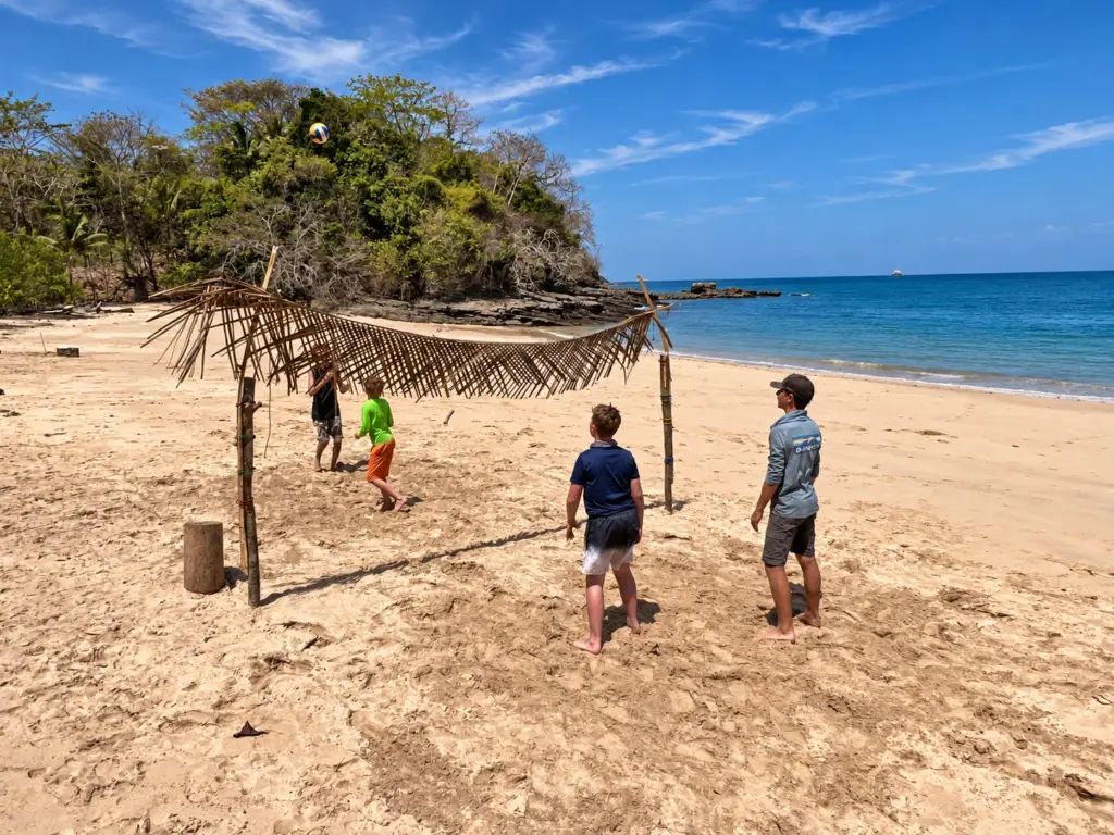 Participants build a palm-roofed shelter frame on a white sand beach