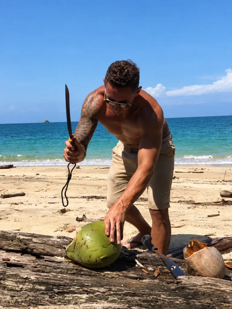 A participant opens a green coconut with a machete on the beach