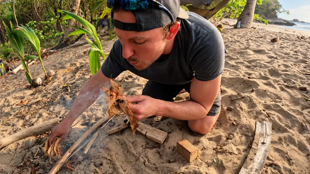 A participant blows embers into a flame while starting a fire on the beach