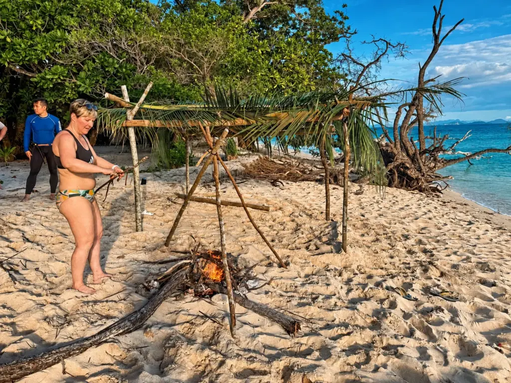 A participant builds a palm shelter frame over a beach fire