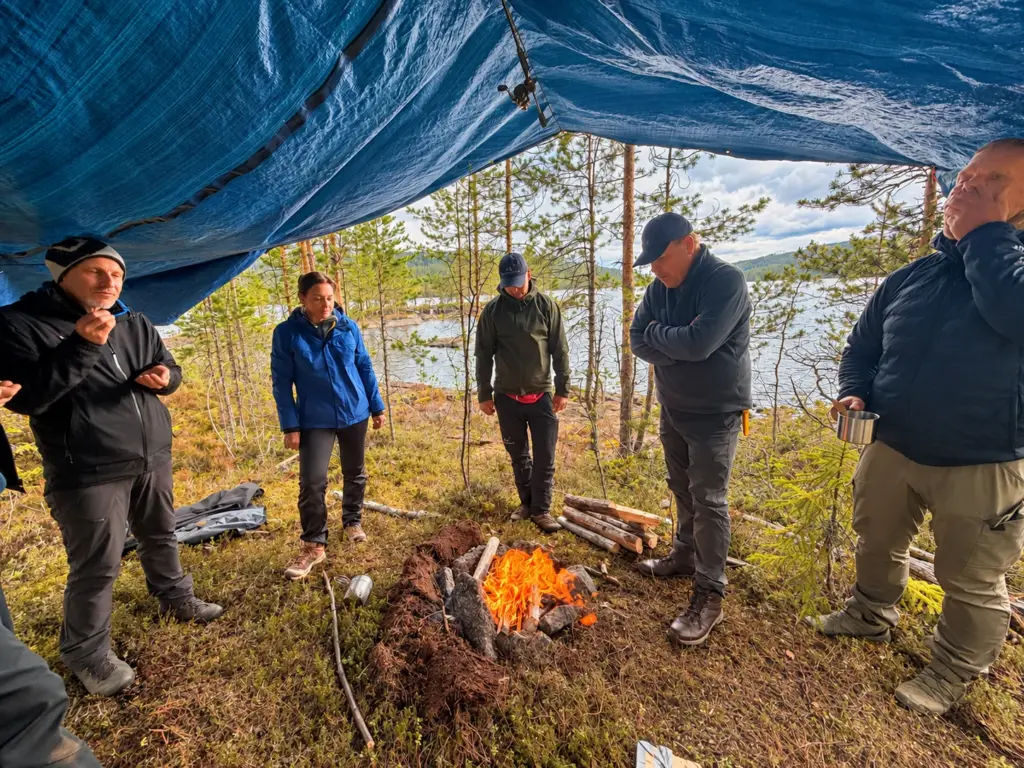 The group gathers around a fire under a tarp at the forest camp