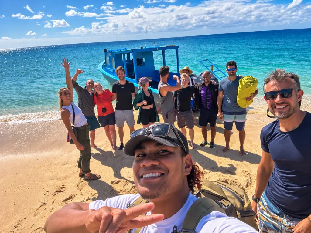 The group waves in a beach selfie beside their boat in Tonga