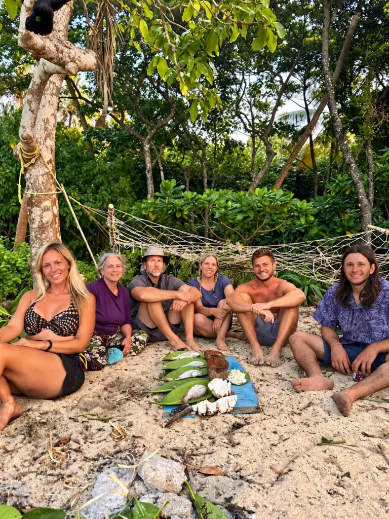 The group sits together on the sand beneath the palms with fresh coconuts