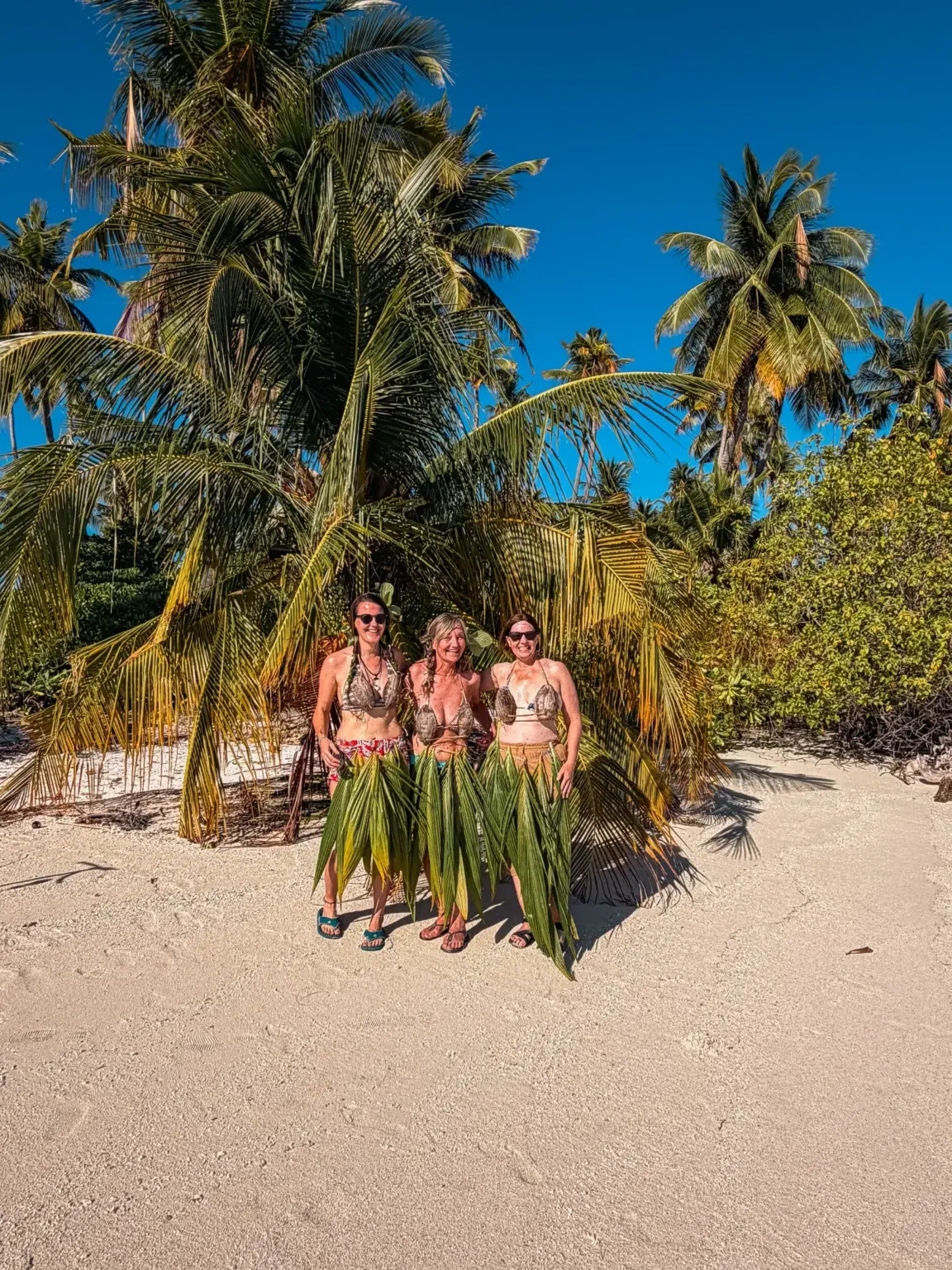 Three women in leaf skirts pose on a Maldives beach. Palm trees, blue sky.