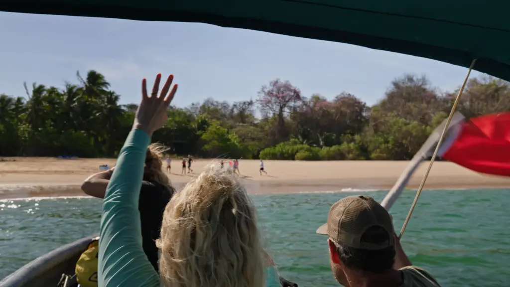 People on a tropical beach seen from a boat, waving towards the shore.