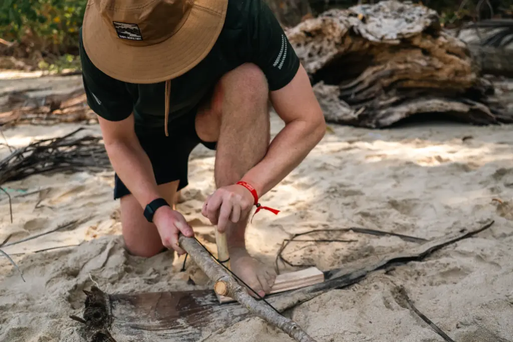 Man on beach using stick and wood for desert island survival training.