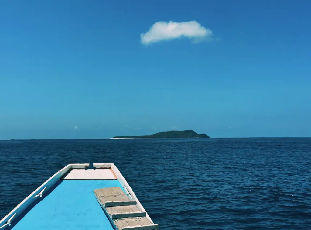Boat heading towards a tropical island under a clear blue sky with a cloud.