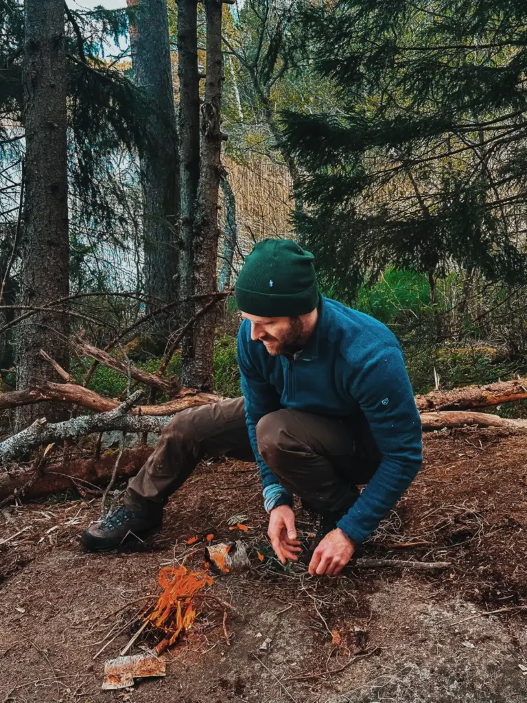 Man building fire in Sweden wilderness, wearing green beanie and blue sweater.