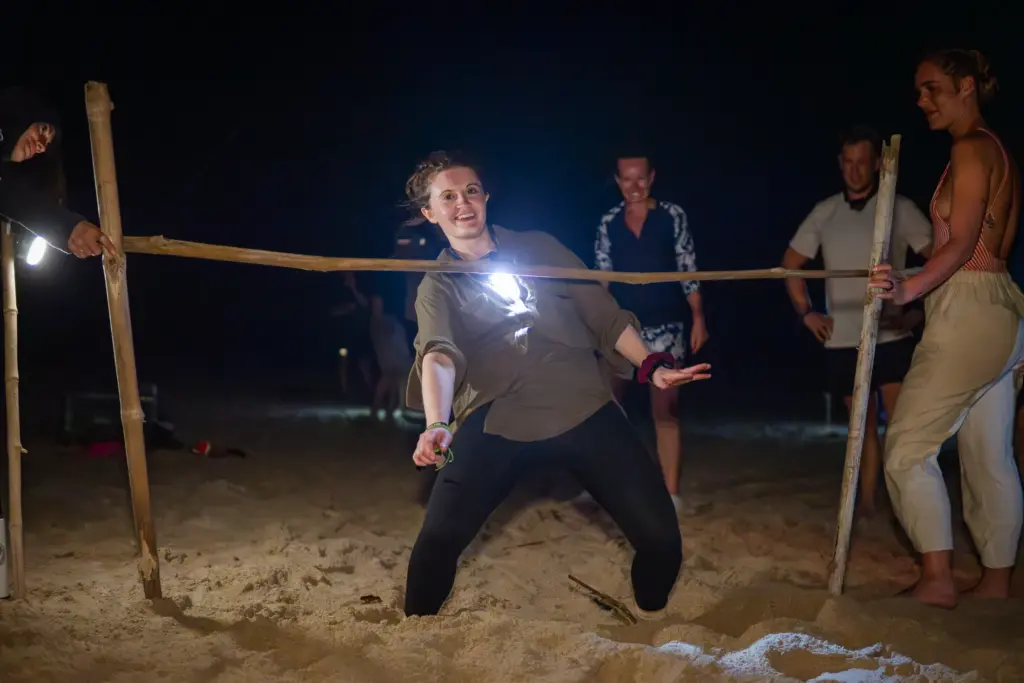Woman bends low under a limbo stick on a sandy beach at night.