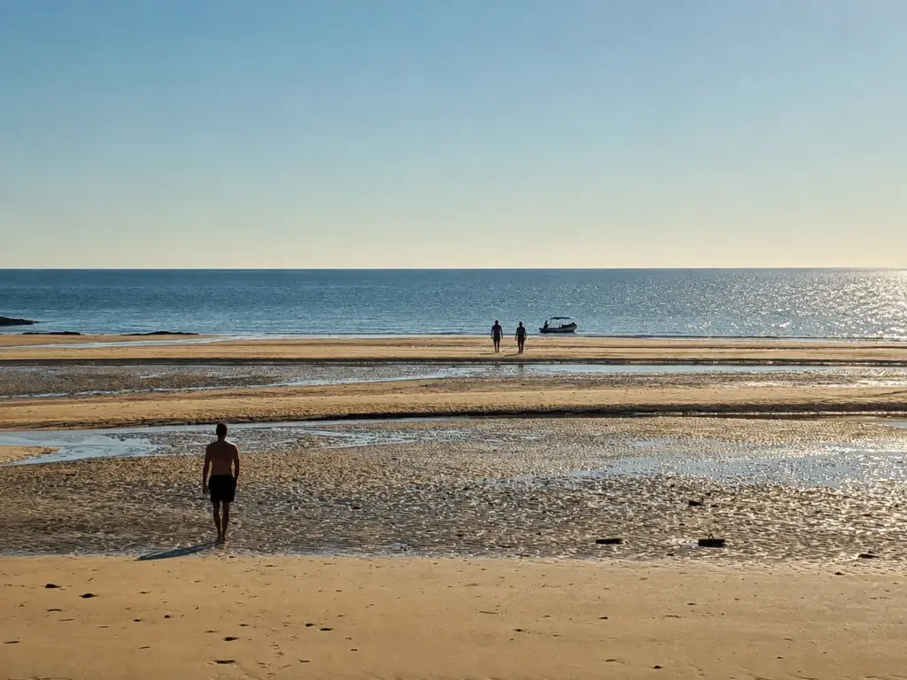 Two people walk on a wet sandbar at low tide, with a third person walking away from the camera.