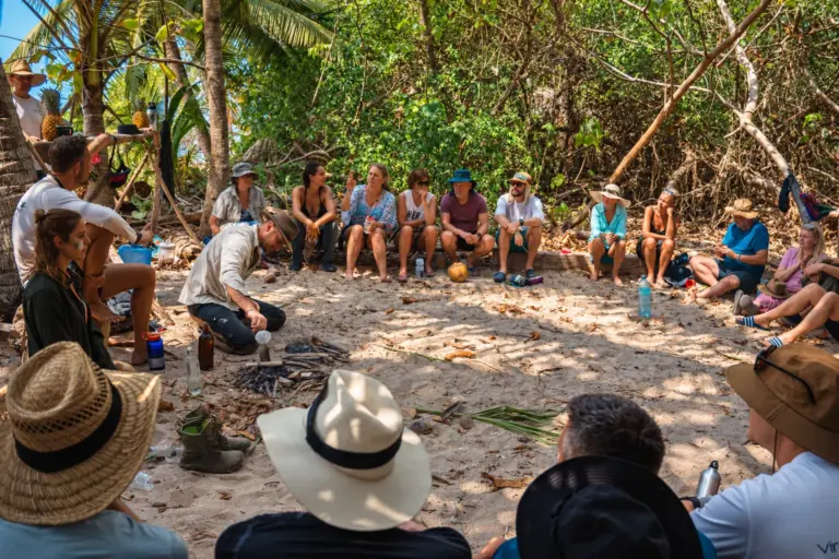 An instructor demonstrates a bushcraft skill to a group seated in a circle on a shaded island beach