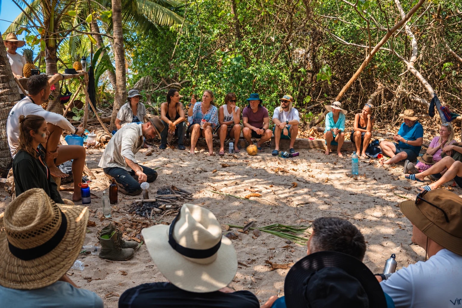 An instructor demonstrates a bushcraft skill to a group seated in a circle on a shaded island beach