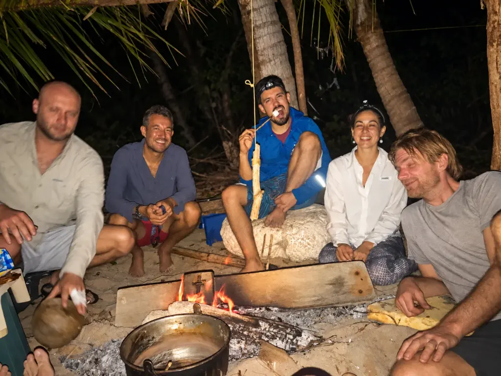 Five expedition members sit around a campfire on a Panama beach at night, cooking and toasting food on sticks